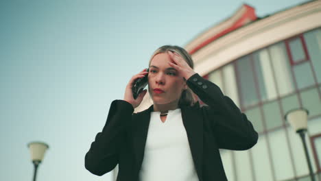 Frustrated girl in professional outfit on phone call looking exhausted and annoyed with building slightly blurred in background and two lampstands visible, expressing visible irritation and fatigue outdoors