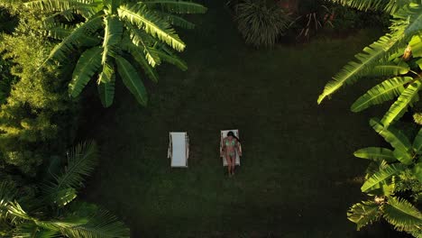 drone shot going down above a young and beautiful woman sitting or laying down in the sunchair in a bikini