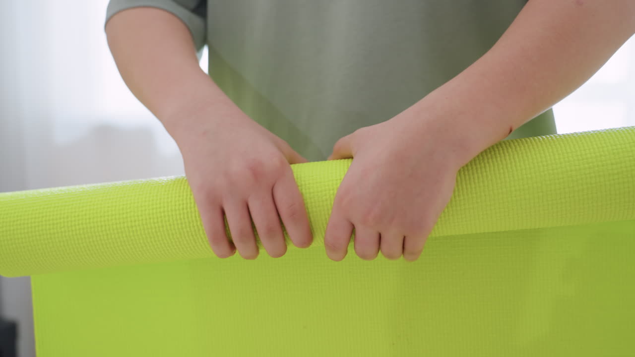 Close up of young boy folding bright green fitness mat in home setting, hands gripping mat firmly with visible arm scratch, reflecting personal responsibility