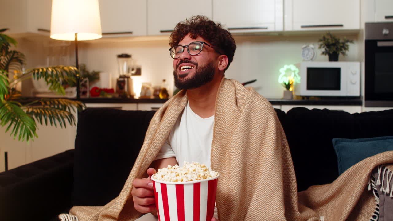 Arabisk mand ser film med popcorn, svøbt i et tæppe, griner og kommenterer på programmet om natten indendørs.