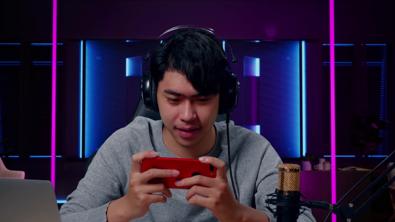 primer plano de un hombre asiático sonriente jugando con auriculares y computadora usando un teléfono móvil jugando en una habitación iluminada por luces de neón en retro