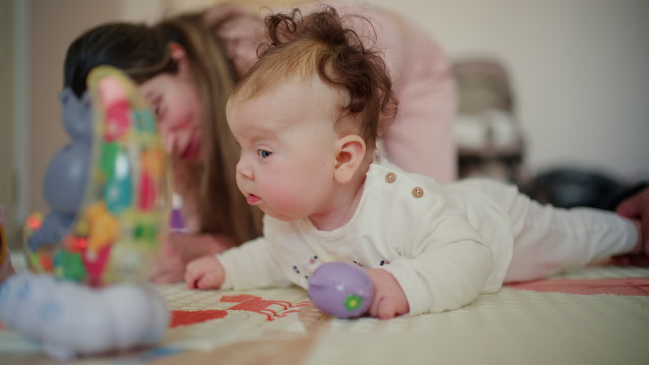 Bebê praticando engatinhar e brincando com brinquedos em um tapete macio, com a mãe supervisionando ao fundo.