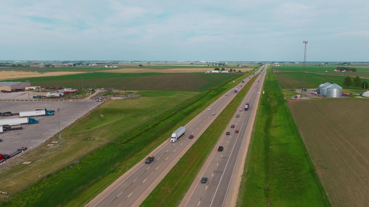 Bisarca commerciale che trasporta veicoli lungo un'autostrada interstatale nell'Iowa rurale, accanto a un grande centro logistico per camion.