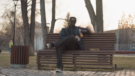 man sits on park bench eating snack and holding drink, sunglasses on, legs crossed, book resting nearby, sunset light, relaxed mood, break in urban park while people stroll in background