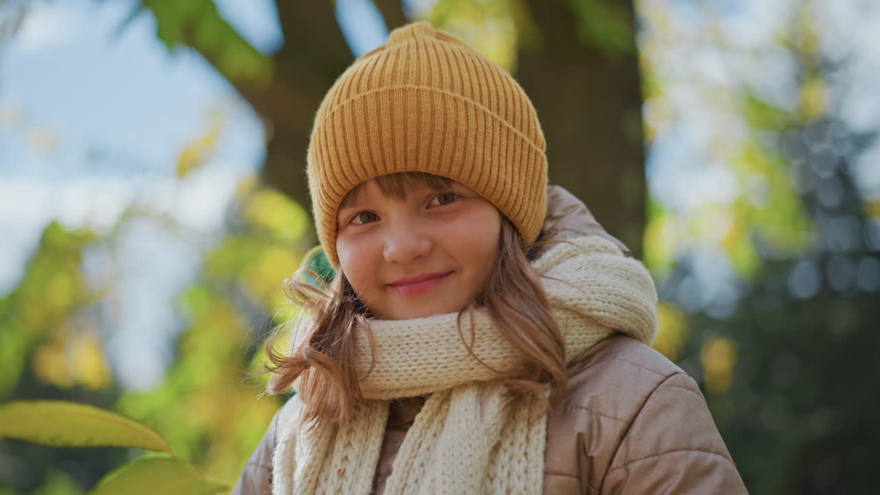 close up of little girl with warm smile holding yellow leaf while standing under tree canopy on leaf strewn park path during crisp autumn day under cloudy sky capturing joyful moment