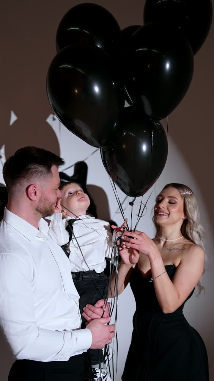Caucasian couple with their little toddler son. Mother is holding several big black balloons. Studio footage. Vertical video.