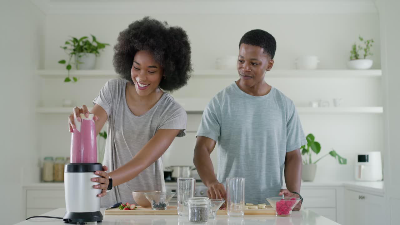 Coppia afroamericana che posiziona la tazza del frullatore tritando frutta per preparare un frullato rosa per due in cucina.