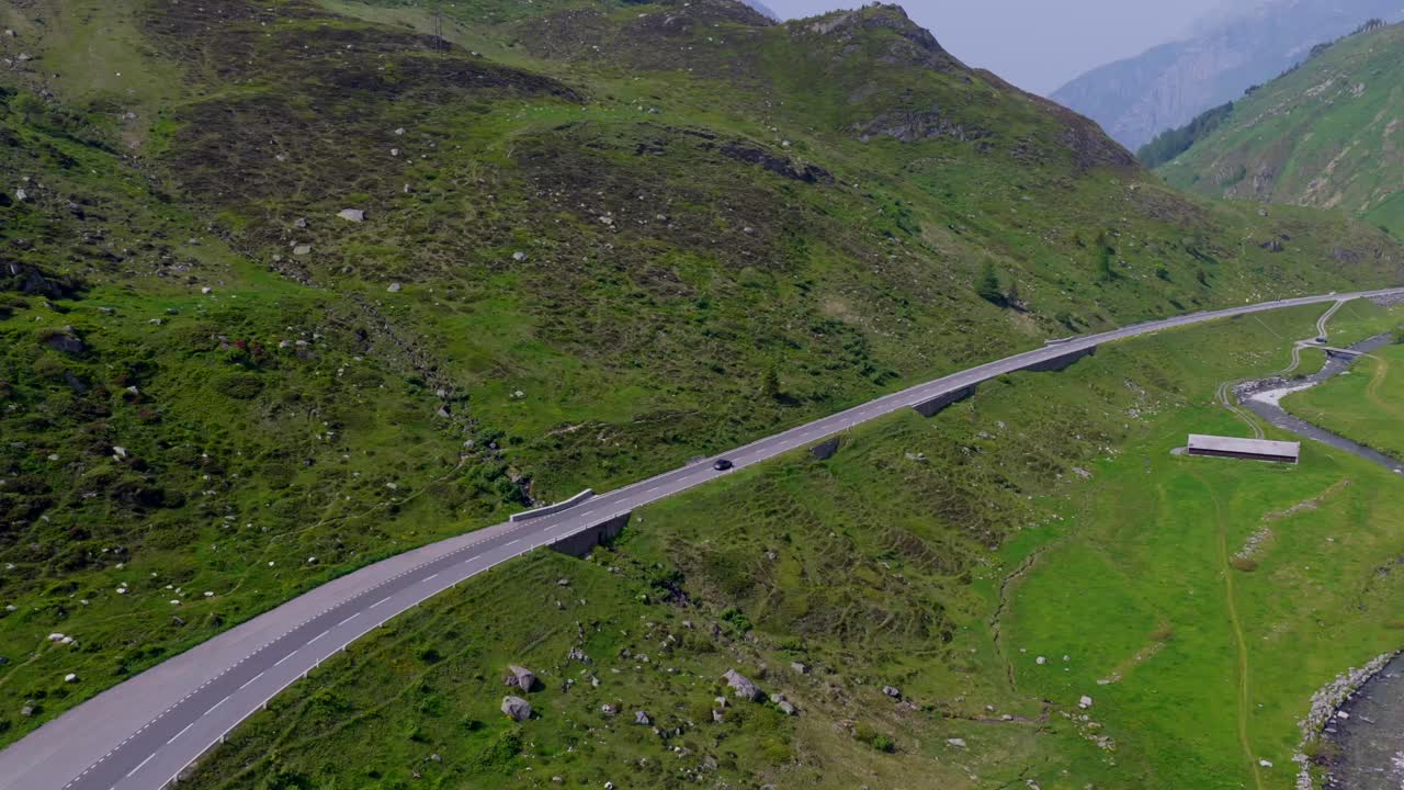 Aerial view of a winding mountain road in Hospental, Switzerland, with cars driving through lush green alpine terrain under clear skies