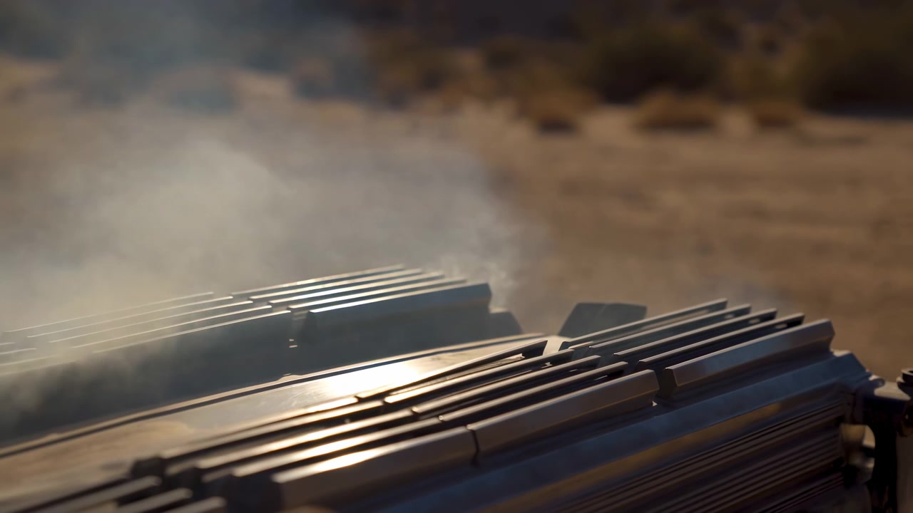 Fumo bianco denso si diffonde costantemente su superfici metalliche a strati mentre la luce del sole colpisce le creste lucide, con messa a fuoco ridotta che separa l'attrezzatura da uno sfondo esterno caldo e asciutto