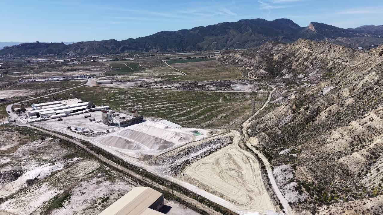Una ripresa con drone in movimento e in rotazione che mostra una grande fabbrica di cemento e un'attività di ghiaia con enormi cumuli di materie prime e magazzini industriali.
