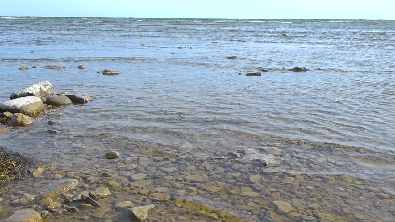 Low angle view of the sea from the shore. Rocky pebble beach with spilling waves and clear water. Slow motion of gentle waves break softly towards shore. Smooth water movement and sun reflection sea