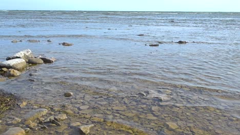 low angle view of the sea from the shore. rocky pebble beach with spilling waves and clear water. slow motion of gentle waves break softly towards shore. smooth water movement and sun reflection sea