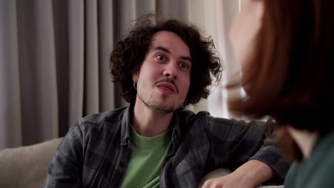 close up over the shoulder a happy brunette guy with curly hair in a mustache and in a checkered gray shirt communicates with his brunette girlfriend while sitting on the sofa at home