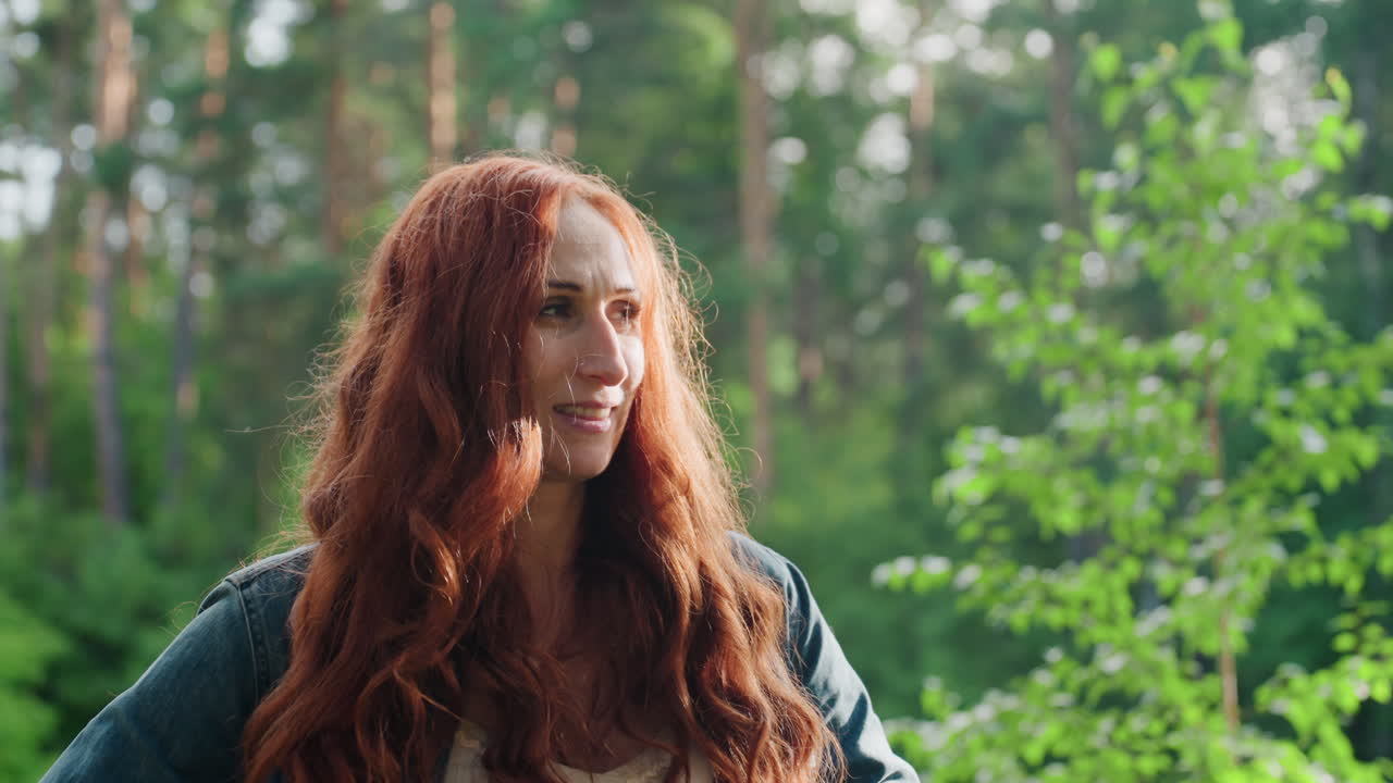 Portrait of beautiful lady with long red hair standing outdoors surrounded by green forest, smiling softly while looking around in warm sunlight, expressing peaceful mood, and natural beauty