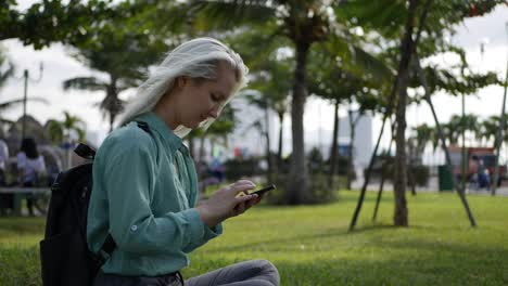 hermosa mujer delgada con el cabello largo rubio en camisa verde se sienta en el suelo y el uso de teléfono inteligente sobre el fondo del parque. niña en la pantalla cuadrada tocando y sonriendo