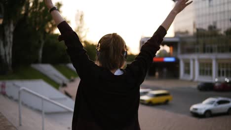 Footage-of-a-stylish-girl-dancing-outdoors-on-the-street-with-modern-building-on-the-background.-Woman-raising-her-hands-with