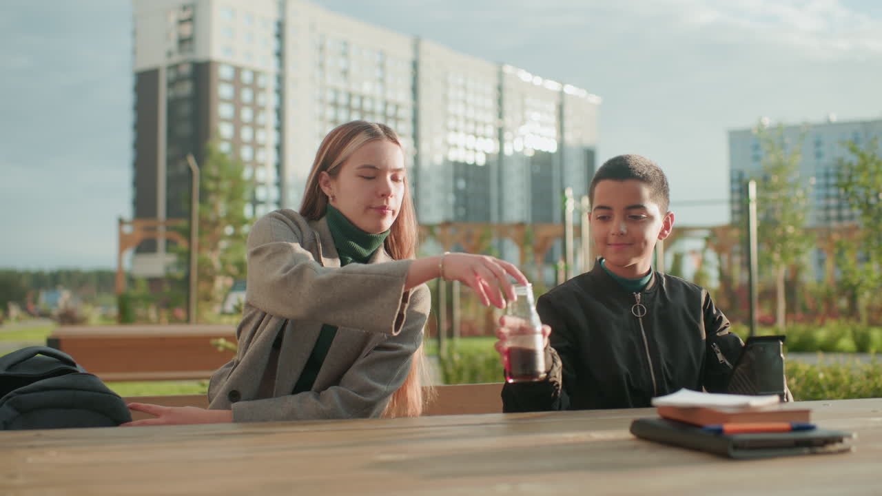 Woman hands over snack to son outdoors as he happily collects it with drink on wooden table, showing warm family connection and joyful bonding moment in bright urban park setting