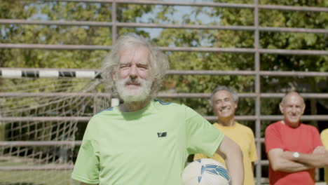 feliz jugador de fútbol senior sosteniendo la pelota y levantando el brazo mientras miraba la cámara en el campo de fútbol