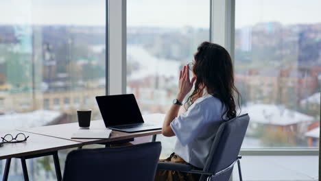young busy lady working at laptop sitting at desk. freelance writer, journalist, copywriter at work. panoramic windows with blurred cityscape at backdrop.