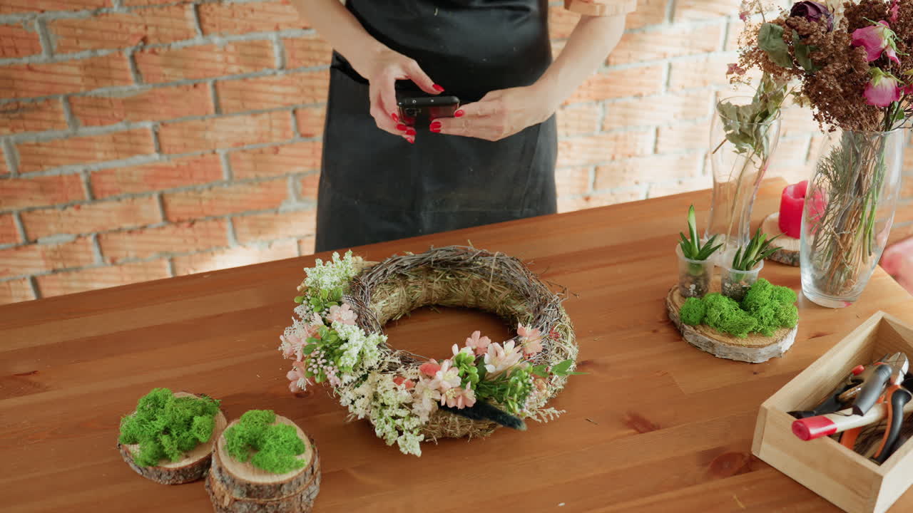 Female florist in black apron arranging floral wreath with pink blossoms and greenery on wooden table, hand reaching toward smartphone beside moss decor, glass vases, and tools in rustic workshop with brick