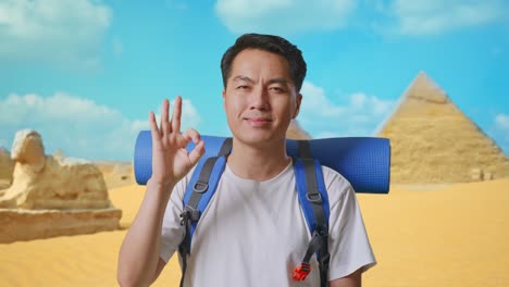 close up of asian male hiker with mountaineering backpack smiling and showing okay gesture to camera while traveling in pyramid of giza