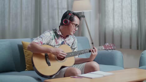 asian man composer with notebook on table wearing headphones and playing guitar at home