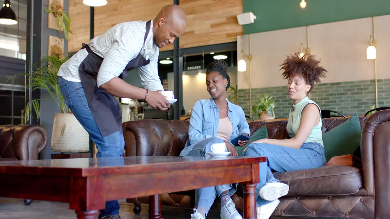 African American server carrying cups or saucers serving drinks on wooden table to women on brown so