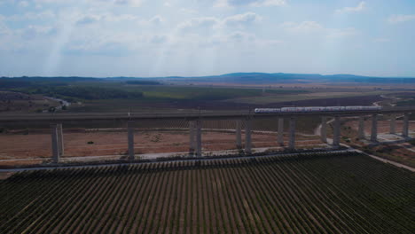 Train-passes-on-a-massive-railway-bridge-over-agricultural-fields-and-vineyards---Colorful-and-cinematic-shot-with-sun-rays-penetrating-from-the-clouds---pull-back-reveal