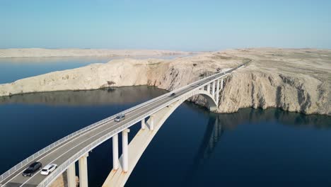 Drone-take-off-over-elegant-arched-bridge-over-blue-Adriatic-and-towards-white-stoney-layers-of-island-Pag-and-lonely-road-meandering