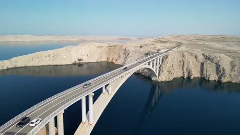 drone take off over elegant arched bridge over blue adriatic and towards white stoney layers of island pag and lonely road meandering