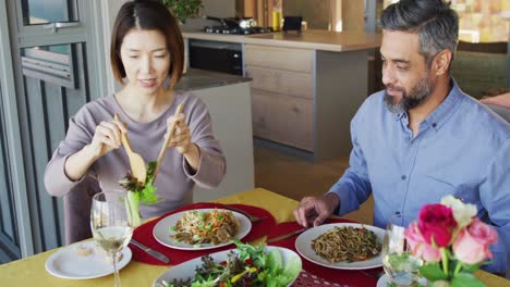 happy diverse couple sitting at table in dining room, eating dinner and drinking wine