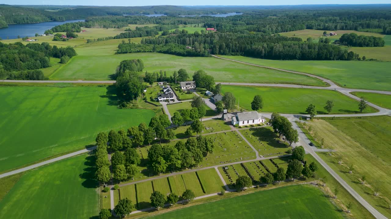 Gunnarsnäsin kirkko, Mellerud, Ruotsi — ilmasta kuvattu parallaksi, valkoinen kirkko, puiden reunustama hautausmaa, laajat vihreät viljelykset, järviä ja metsäinen horisontti näkyvissä, aurinkoinen kesäpäivä.