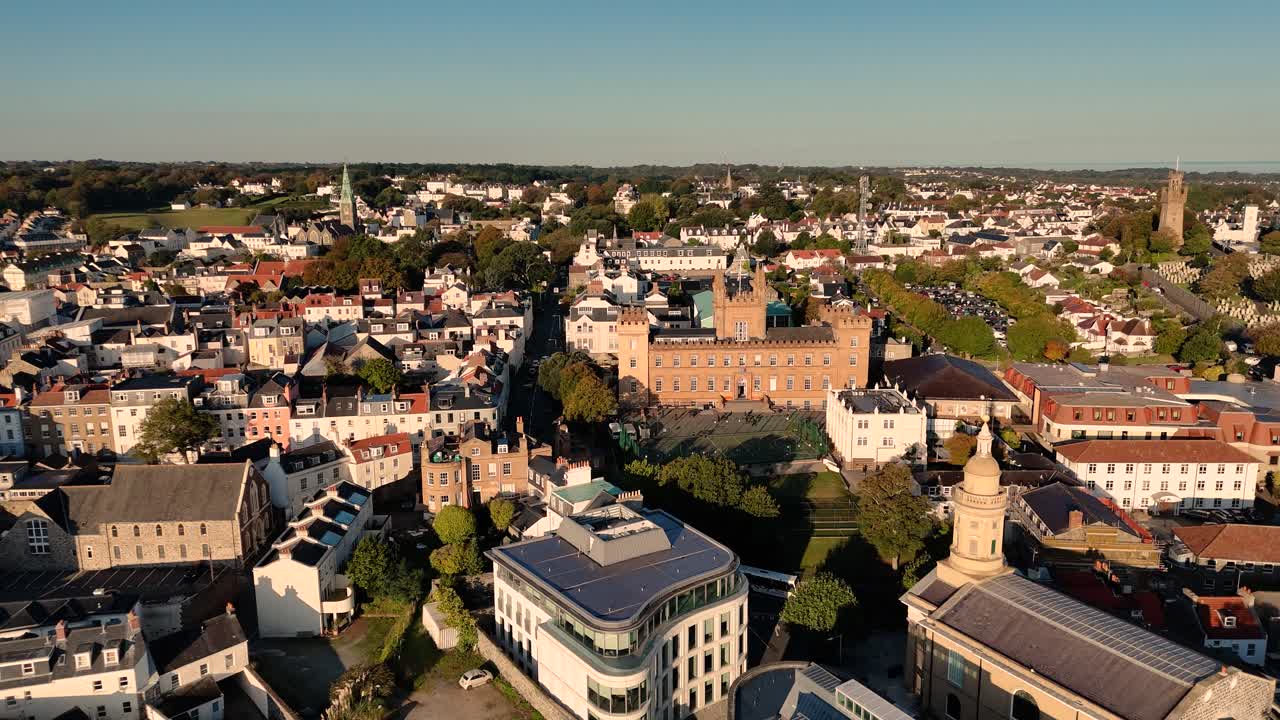 St Peter Port Guernsey: Sakte, lavt droneopptak over Royal Court, St James og hustak mot Elizabeth College, med utsikt over horisonten og hele øya på en skyfri sommerdag i sterk sol.