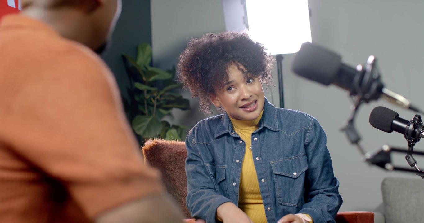 Afroamerikansk kvinde i denim svarer på gestus fra vært til venstre på settet med mikrofoner, mens hun taler.