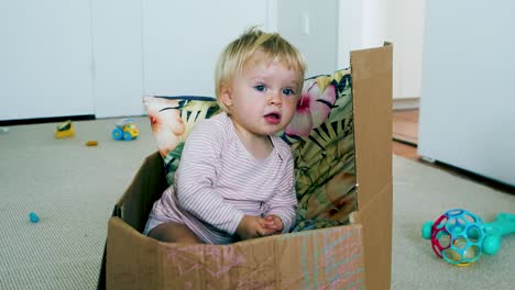 Small-Toddler-sitting-inside-a-cardboard-boxing-filled-with-pillows,-rocking-back-and-forward