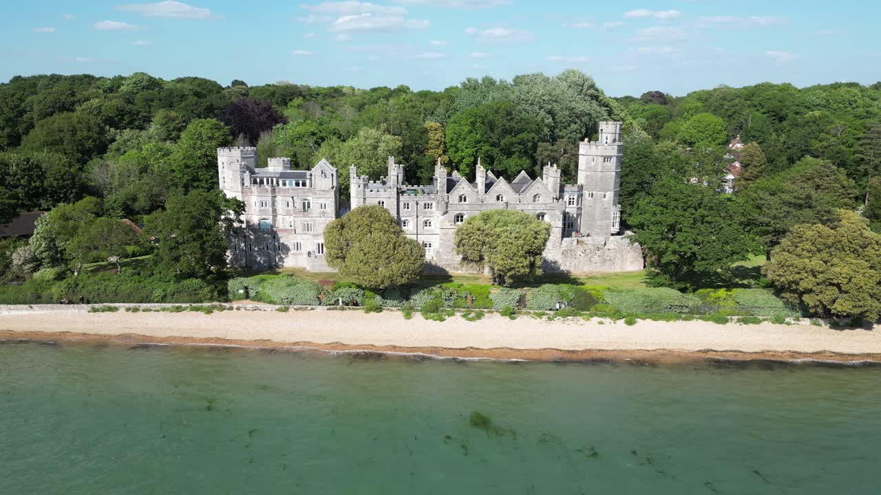 An impressive historic castle stands majestically on a sandy beach, bordered by a dense forest and calm waters under a bright sky.