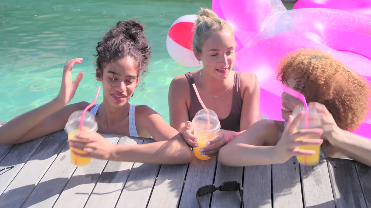 Amiche di diverse etnie sorseggiano bevande all'arancia con cannucce rosa e fanno tintinnare i bicchieri a bordo piscina dopo una partita a palla.