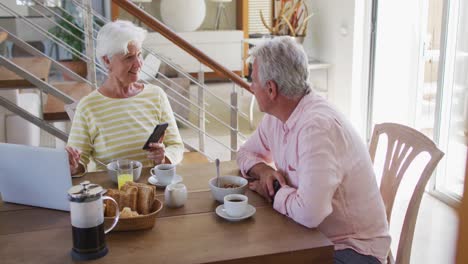 senior caucasian couple talking to each other using smartphone and laptop having breakfast together