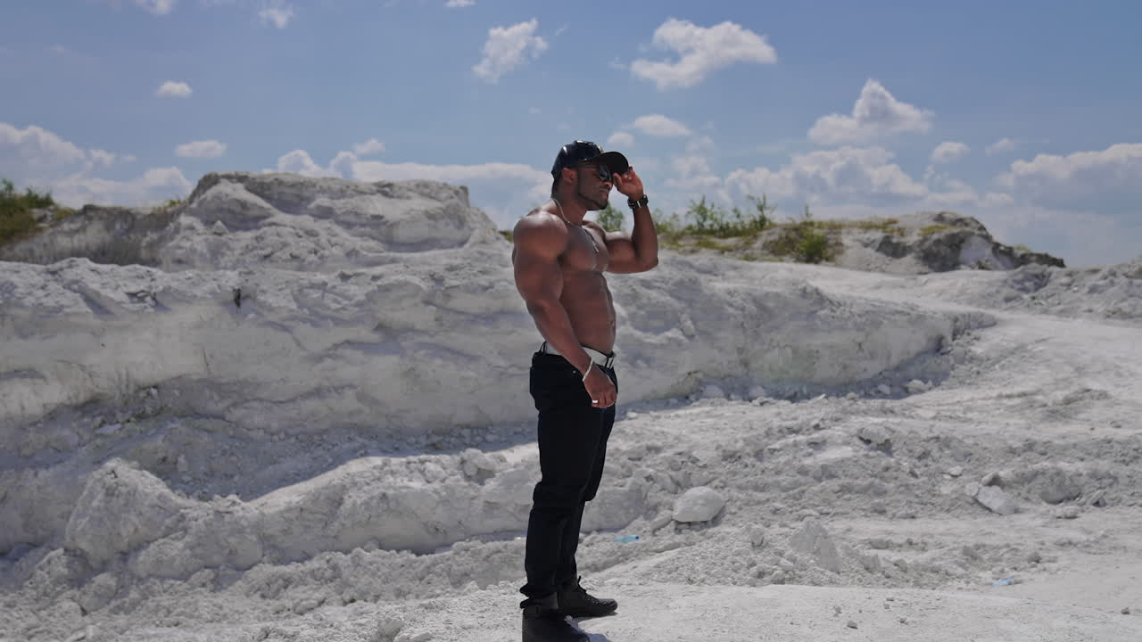 Handsome guy without shirt standing on white rocky background. Dark skinned guy in black cap and glasses with muscular body outdoors. Profile view.