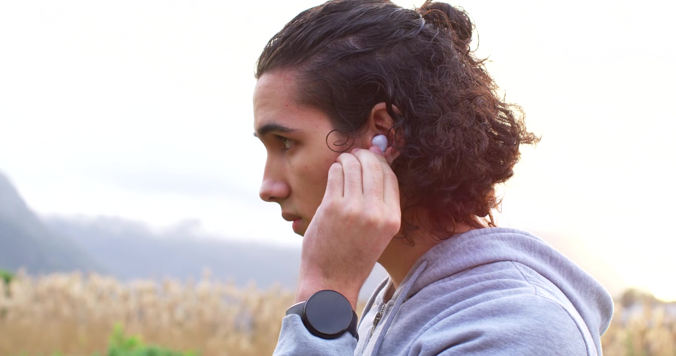 Hombre afroamericano con sudadera con capucha insertando un auricular blanco en su oreja derecha, sobre un fondo liso con espacio para texto.