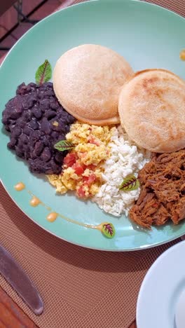 latin american breakfast with cornmeal arepas, grated white cheese, shredded beef, black beans and eggs perico. traditional decorated dish on wooden table