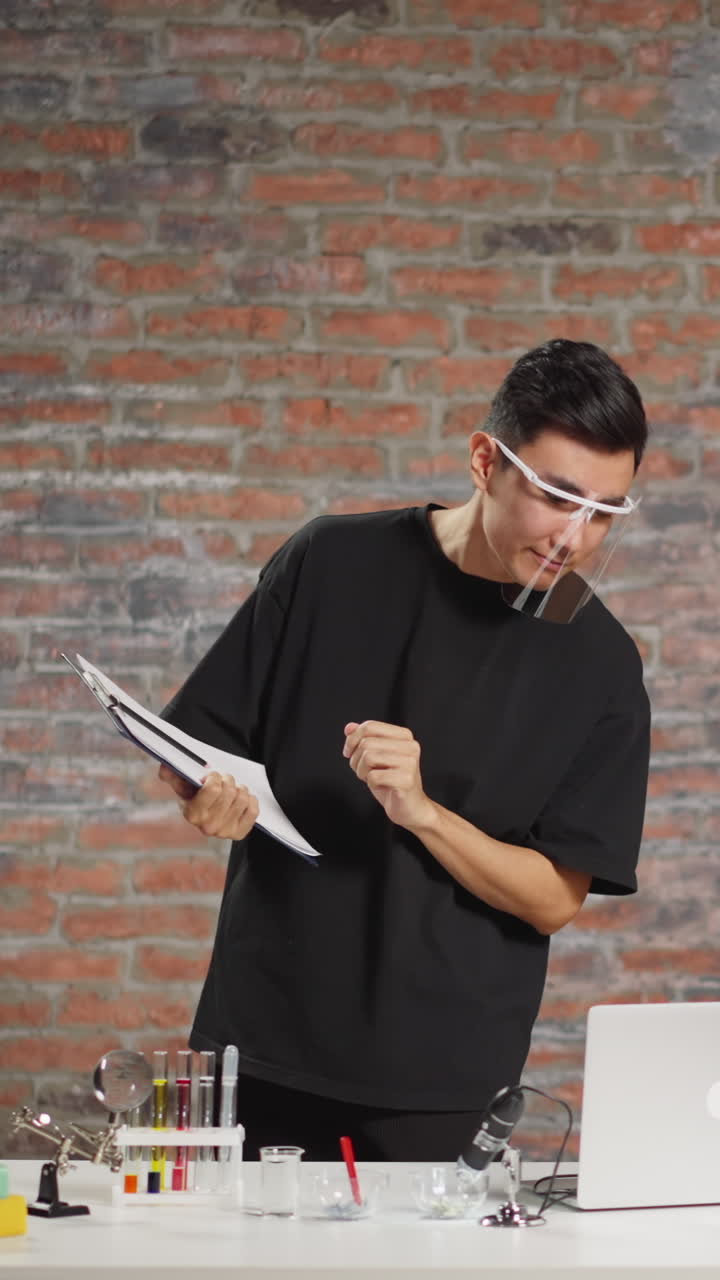 Young Asian man with plastic face shield and clipboard does scientific experiment and records video for blog with laptop in home laboratory