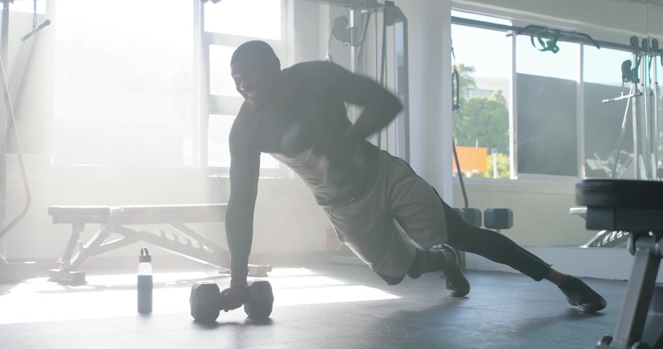 Uomo afroamericano in posizione di plank che inizia delle remate, ruotando per fare un piegamento con manubri per il core in palestra.