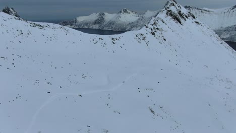 Drone-view-in-Tromso-area-in-winter-flying-over-a-snowy-mountain-in-Segla,-Norway-with-the-sea-on-the-horizon