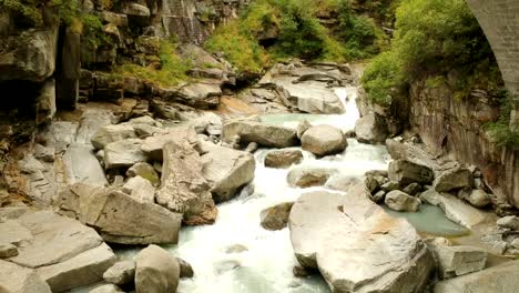 Slow-aerial-dolly-shot-under-a-bridge-and-over-a-rocky-alpine-stream-with-glacial-water-crashing-over-lightly-coloured,-heavily-eroded-rocks