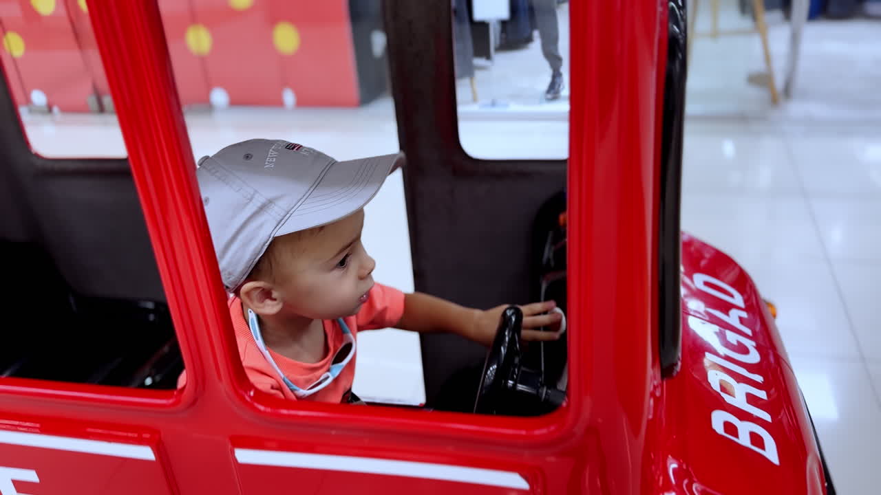 Adorable toddler in a cap sits in the moving toy fire car. Excited kid jumps up, ogresses buttons and turns the wheel. Close up.