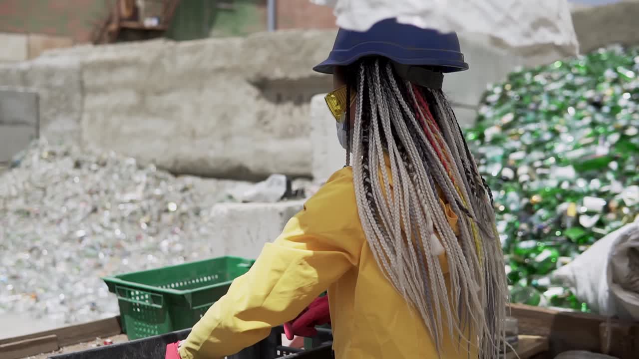 Young woman in hard hat standing against the pile of broken glass, used bottles next to the wall. Girl in yellow jacket crashing old glass bottles for further recycling. Rare view. Close up