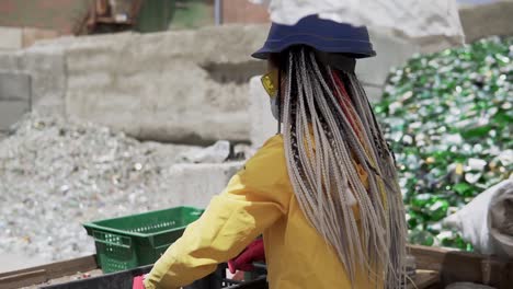 young woman in hard hat standing against the pile of broken glass, used bottles next to the wall. girl in yellow jacket crashing old glass bottles for further recycling. rare view. close up