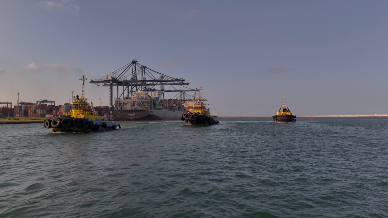 Three tugboats on the water in front of a busy port terminal, with cargo ships and cranes in the background under clear daytime skies.