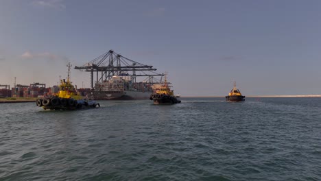 three tugboats on the water in front of a busy port terminal, with cargo ships and cranes in the background under clear daytime skies.